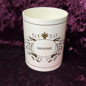 May include: A cylindrical container with a cream-colored exterior and a brown decorative design. The design includes the word "GOSSYPION" within an ornate frame. The container has a simple, open top.