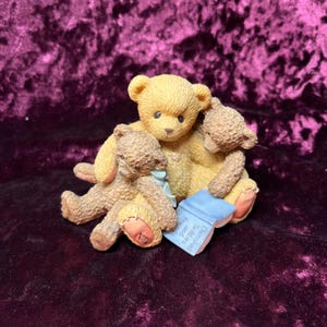 May include: A figurine of three teddy bears in various shades of brown and tan. One bear holds a blue book with the words "Cherished Teddies and Friends". The bears are clustered together, creating a heartwarming scene.