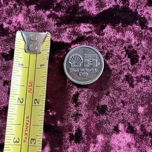 May include: A silver-colored metal token with the Shell Motorist Club logo. The logo features a stylized seashell and a lighthouse. The token is about 1 inch in diameter.