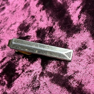 Puede incluir: Un pasador de corbata plateado con un diseño geométrico en un extremo. El pasador tiene forma rectangular con un aspecto ligeramente desgastado, descansando sobre una superficie de terciopelo morado oscuro. El diseño presenta un triángulo con un patrón de pequeños círculos.