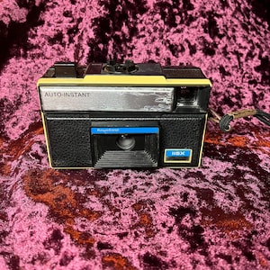 May include: A vintage Keystone 115X Auto-Instant camera. The camera is black with yellow and silver accents. The camera has a blue rectangular frame around the lens and the text "AUTO-INSTANT" is printed on the top.