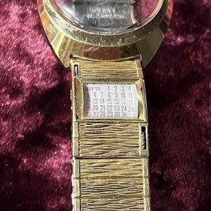 May include: Gold-toned vintage wristwatch with a clear face and a calendar display for May '74. The watch features a textured band and a rectangular calendar window with the days of the month.