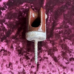 Pode incluir: Um isolador elétrico de cerâmica esmaltada castanha com uma ranhura superior em forma de U. A base é composta por um material cinzento texturado e inclui um longo parafuso metálico roscado virado para baixo para fixação.