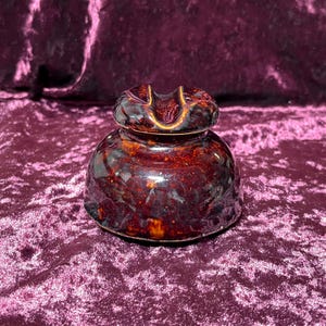 May include: A vintage, brown ceramic insulator with a glossy finish. The insulator has a rounded base and a top with a U-shaped groove. The ceramic has a mottled appearance with shades of brown and reddish-orange.