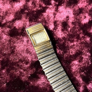 May include: Vintage digital watch with a gold-tone rectangular face and a silver-tone expanding band. The watch face displays numbers. The watch is set against a dark red velvet background.