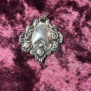 May include: A silver pendant with an ornate design. The pendant features a central, smooth area surrounded by detailed floral and face-like motifs. It has a loop at the top for a chain. The background is a deep burgundy velvet.