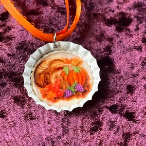 May include: A circular pendant with a harvest design, showing a pumpkin, grapes, and leaves. The pendant is set in a white bottle cap and hangs from an orange ribbon. The background is a textured, dark pink fabric.