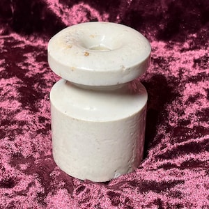 Puede incluir: Un tapón de botella de cerámica de color claro, con una base cilíndrica y una parte superior circular con un agujero en el centro. El tapón tiene una superficie lisa, ligeramente texturizada y una paleta de colores neutros.
