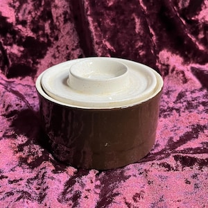 May include: A brown ceramic butter dish with a white lid. The lid has a recessed center. The dish is cylindrical and sits on a dark purple velvet surface. The butter dish is a kitchenware item.