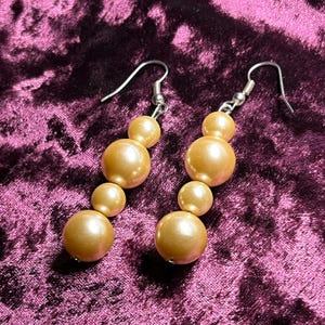 Pode incluir: Um par de brincos pendurados com uma série de contas redondas graduadas, cor de pêssego. Os brincos têm ganchos de orelha prateados e são exibidos contra um fundo de veludo roxo escuro.