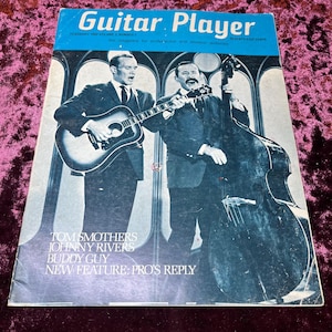 以下が含まれることがあります： トム・スマザーズとジョニー・リバーズをフィーチャーした、ヴィンテージの「ギタープレイヤー」誌の表紙。表紙には雑誌のタイトルと号の詳細が記載された青いバナーがあります。画像には、ギターを持ったスーツ姿の男性が2人写っています。雑誌にはバディ・ガイも掲載されています。