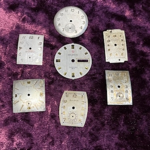 May include: A collection of six vintage watch dials in various shapes including round, square, and rectangular. The dials feature metallic silver and gold tones with printed numerals, indices, and brand names such as Helbros, Bulova, and Elgin.