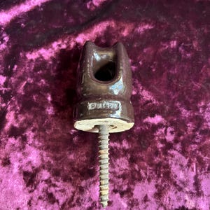 May include: A dark brown glazed ceramic electrical insulator featuring a U-shaped top groove and a threaded metal screw extending from the base. The ceramic body displays the embossed text "pp1929" on its front surface, with a smooth, reflective finish.