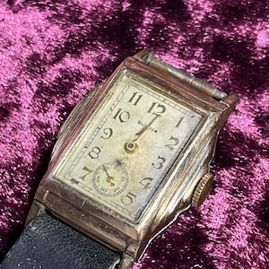 May include: Vintage Bulova wristwatch with a rectangular gold-toned case and a cream-colored face. The watch features black numerals, a small seconds dial, and a black leather band. The watch face displays the Bulova brand name.