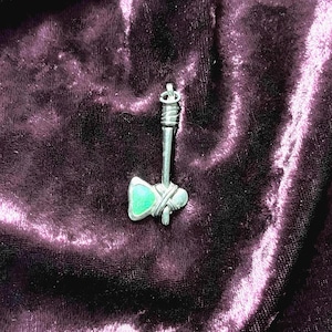 May include: A silver axe pendant with a green stone inset. The pendant is on a dark purple velvet background.