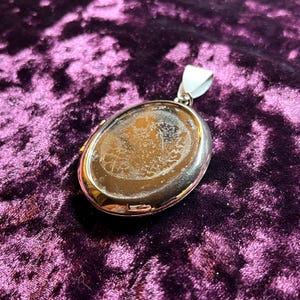 Könnte beinhalten: Ovale silberne Medaille mit leerem Innenraum, bereit für ein Foto oder Andenken. Das Medaillon hat eine polierte Silberoberfläche und eine kleine Schlaufe für eine Kette. Der Hintergrund ist tiefvioletter Samt.