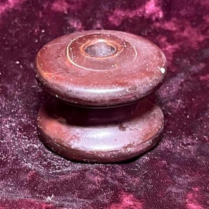 May include: A dark red, spool-shaped object with a central hole. The object has a slightly worn appearance, with visible imperfections and a glossy finish. The design features two rounded, disc-like sections connected by a narrower, cylindrical portion.