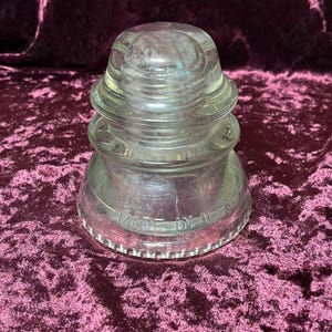 Op de afbeelding: Transparante glazen isolator met een afgeronde bovenkant en een brede basis. De isolator heeft een geribbeld ontwerp en de tekst "MADE IN U.S.A." is zichtbaar. Het object staat op een bordeauxrode ondergrond.