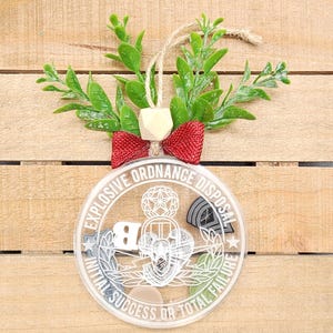 Puede incluir: Un adorno navideño redondo transparente con un diseño blanco de una insignia de Desactivación de Artefactos Explosivos (EOD). El adorno tiene el texto "Explosive Ordnance Disposal" y "Initial Success or Total Failure" alrededor del borde. El adorno está colgado de un lazo rojo y una cuerda.