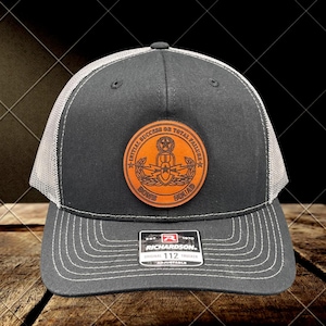 以下が含まれることがあります： 白メッシュの背面と革製パッチが付いた、濃いグレーのトラッカーハット。パッチには「Initial Success or Total Failure」と「Bomb Squad」の文字が書かれています。帽子にはRichardson 112のタグが付いています。