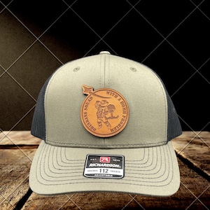 May include: Olive green and black trucker hat with a leather patch that reads "Drinking Squad With a Bomb Problem." The hat has a curved brim and a mesh back. The Richardson brand label is visible.