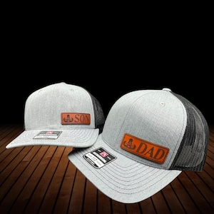 以下が含まれることがあります： 黒いメッシュの背面が付いた2つのグレーのトラッカーハット。1つの帽子には「SON」と書かれた茶色の革パッチがあり、もう1つは「DAD」と書かれています。どちらの帽子にもRichardson 112のタグが付いています。