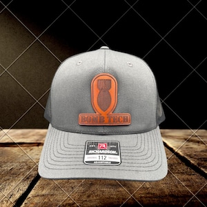 May include: Grey trucker hat with a brown leather patch featuring a bomb design and the text "BOMB TECH". The hat has a mesh back and a curved peak. The Richardson brand label is visible.