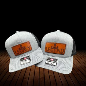 May include: Two grey trucker caps with black mesh backs. Each cap features a brown leather patch with the words "LEGACY" and "LEGEND" in embossed lettering. The caps have a Richardson 112 adjustable tag.