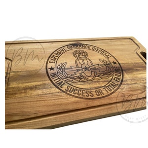 Puede incluir: Una tabla de cortar de madera con un diseño grabado con láser del emblema de la Desactivación de Artefactos Explosivos (EOD). El emblema presenta una bomba con rayos y una corona de hojas. El texto alrededor del emblema dice "Explosive Ordnance Disposal Initial Success or Total Failure".