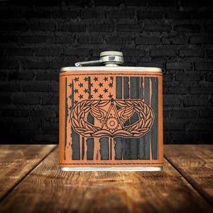 May include: A rectangular, stainless steel flask with a brown leather cover. The leather is etched with an American flag design, including stars and stripes, and a military emblem. The flask has a silver cap and a hinged spout.