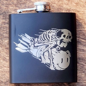 以下が含まれることがあります： 銀色のキャップと、翼のある爆弾に乗った骸骨のレーザー彫刻が施された黒い金属製のフラスコです。