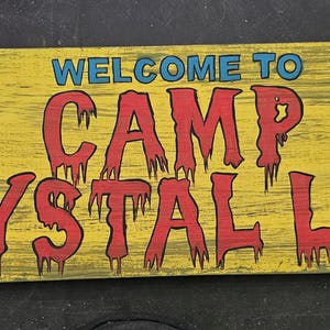 May include: A rectangular wooden sign with a yellow background and the words "WELCOME TO CAMP CRYSTAL LAKE" in red and blue lettering. The sign has a distressed, aged appearance.