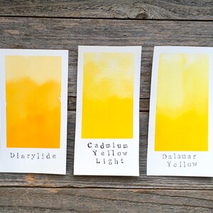 May include: Three rectangular white paper swatches featuring watercolor paint gradients in shades of yellow. The swatches are labeled with black text reading Diarylide, Cadmium Yellow Light, and Dalamar Yellow, showing distinct pigment variations.