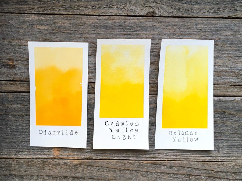 May include: Three rectangular white paper swatches featuring watercolor paint gradients in shades of yellow. The swatches are labeled with black text reading Diarylide, Cadmium Yellow Light, and Dalamar Yellow, showing distinct pigment variations.