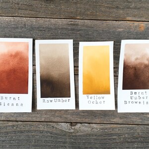 May include: Four rectangular white paper swatches featuring watercolor gradients. The labels read Burnt Sienna, Raw Umber, Yellow Ocher, and Burnt Umber Brownish. Each swatch displays a distinct color transition from light to dark tones.