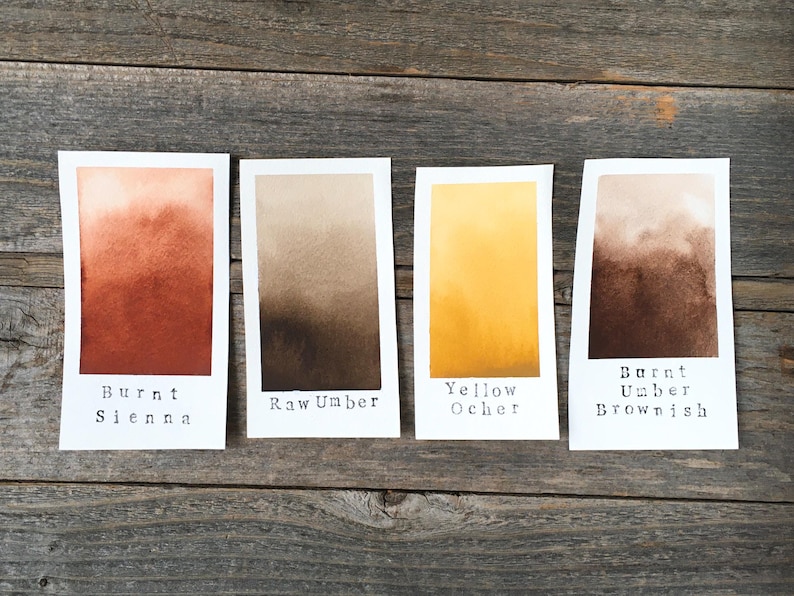 May include: Four rectangular white paper swatches featuring watercolor gradients. The labels read Burnt Sienna, Raw Umber, Yellow Ocher, and Burnt Umber Brownish. Each swatch displays a distinct color transition from light to dark tones.