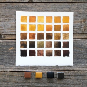 May include: A square sheet of white paper featuring a grid of thirty hand-painted watercolor swatches in various shades of yellow, ochre, brown, and black. Below the paper are four individual square watercolor pans in brown, yellow, and black.