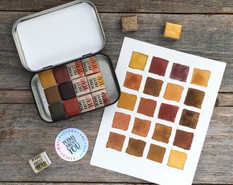 May include: A metal tin containing twelve watercolor paint pans labeled Poems About You, a white paper swatch card with twenty-four painted squares in shades of brown, gold, and red, two loose paint pans, and a circular sticker with the text Poems About You.