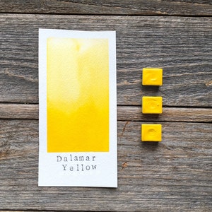 May include: A rectangular white paper swatch featuring a vibrant yellow watercolor wash with the text "Dalamar Yellow" printed at the bottom, accompanied by three small, square, solid yellow watercolor paint pans arranged in a vertical column.
