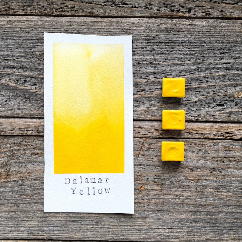May include: A rectangular white paper swatch featuring a vibrant yellow watercolor wash with the text "Dalamar Yellow" printed at the bottom, accompanied by three small, square, solid yellow watercolor paint pans arranged in a vertical column.
