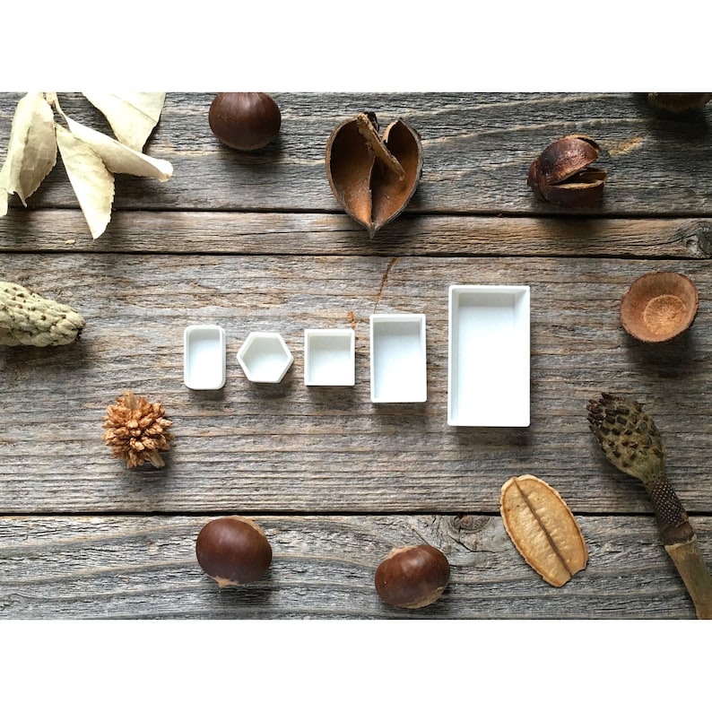 Może przedstawiać: Sześć białych palet do farb akwarelowych w r&oacute;żnych rozmiarach i kształtach ułożonych w rzędzie na drewnianej powierzchni. Palety są puste i gotowe do użycia.