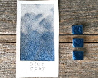 Può includere: Un campione di carta rettangolare con una sfumatura ad acquerello dal bianco al blu-grigio profondo, con il testo "Blue Gray" stampato in basso. Tre piccoli godet quadrati di acquerello blu metallizzato sono posizionati a destra.