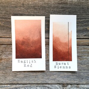 May include: Two watercolor paint swatches on white cardstock. The left swatch is labeled "English Red" and is a reddish-brown color. The right swatch is labeled "Burnt Sienna" and is a darker brown color.