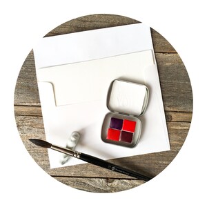 Puede incluir: Una peque&ntilde;a caja de metal plateado que contiene cuatro pastillas de acuarela cuadradas en tonos morado oscuro y rojo brillante, acompa&ntilde;ada de un pincel de mango negro y un sobre blanco con una tarjeta en blanco.