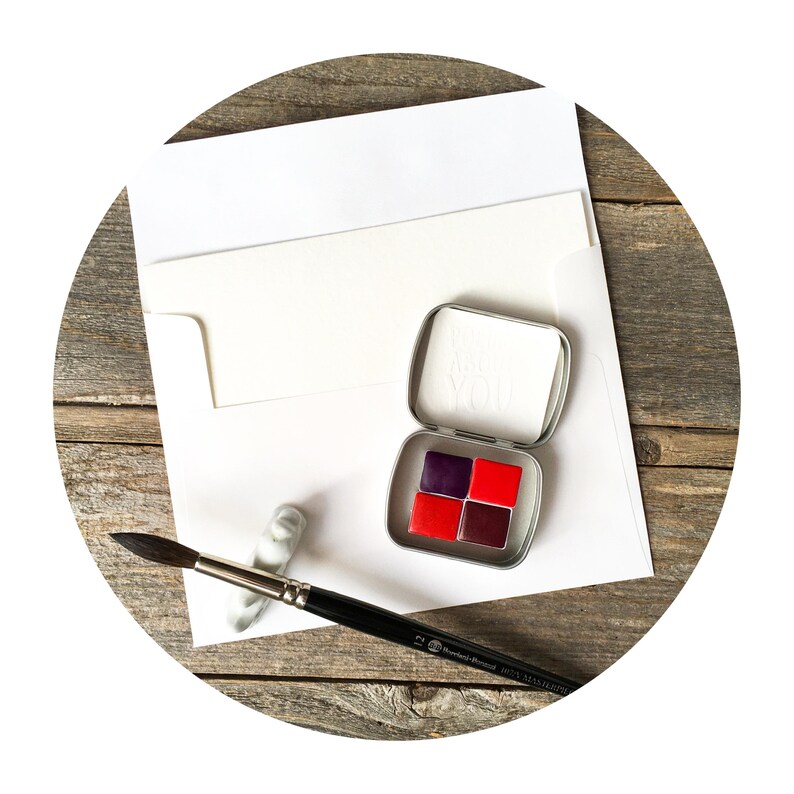 Puede incluir: Una peque&ntilde;a caja de metal plateado que contiene cuatro pastillas de acuarela cuadradas en tonos morado oscuro y rojo brillante, acompa&ntilde;ada de un pincel de mango negro y un sobre blanco con una tarjeta en blanco.