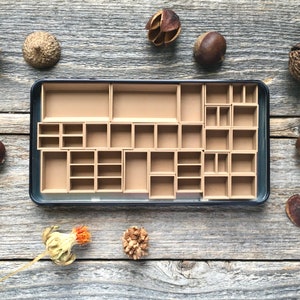 Może przedstawiać: Czarne metalowe pudełko z wyjmowaną tacką zawierającą 24 małe przegr&oacute;dki. Przegr&oacute;dki są wykonane z brązowego kartonu i są puste. Pudełko jest otoczone r&oacute;żnymi naturalnymi przedmiotami, w tym żołędziami, kasztanami i strąkami nasion.