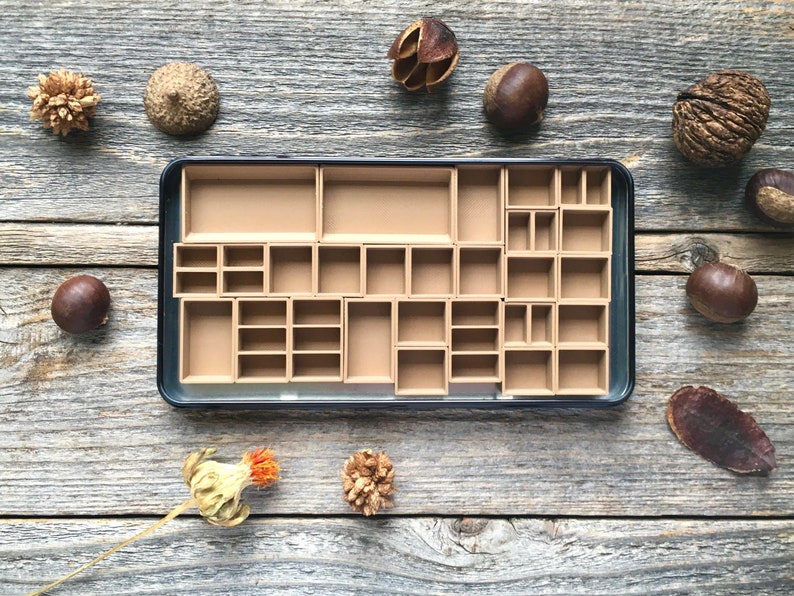 Może przedstawiać: Czarne metalowe pudełko z wyjmowaną tacką zawierającą 24 małe przegr&oacute;dki. Przegr&oacute;dki są wykonane z brązowego kartonu i są puste. Pudełko jest otoczone r&oacute;żnymi naturalnymi przedmiotami, w tym żołędziami, kasztanami i strąkami nasion.