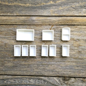 May include: A set of ten white watercolour paint pans, arranged in a row on a wooden surface. The pans are rectangular and have a slightly raised edge.