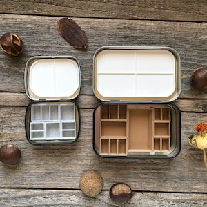 Może przedstawiać: Dwie otwarte metalowe palety do farb z wyjmowanymi tackami. G&oacute;rna paleta ma białą tackę z czterema przegr&oacute;dkami. Dolna paleta ma brązową tackę z 12 przegr&oacute;dkami.