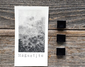 Könnte beinhalten: Ein rechteckiges weißes Papiermuster mit einem schwarz-grauen Aquarellverlauf und dem aufgedruckten Wort Magnetite am unteren Rand, begleitet von drei kleinen, quadratischen, soliden schwarzen Aquarellnäpfchen in einer vertikalen Reihe.